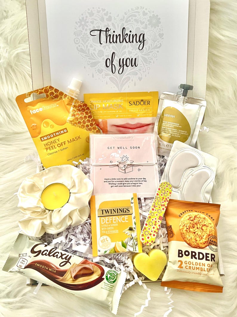 May include: A gift box filled with self-care items, including a face mask, lip mask, eye patches, tea, chocolate bar, and snacks. The box is decorated with the phrase "Thinking of you" and includes a scrunchie and a heart-shaped soap.