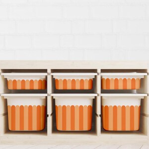 Pode incluir: Uma unidade de armazenamento de madeira com seis compartimentos, cada um contendo um recipiente listrado branco e laranja. Os recipientes têm um design ondulado na parte superior e são dispostos em duas fileiras de três. A unidade está contra uma parede de tijolos brancos.
