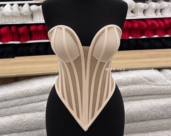 May include: A beige strapless corset top featuring a sweetheart neckline, vertical boning channels, and sheer mesh panels. The garment has a pointed hemline and includes the text WITH YOU printed in white on the right side of the bodice.
