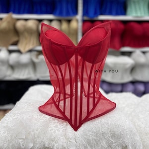 May include: A red corset top featuring a sheer mesh bodice with vertical boning, a sweetheart neckline, and a pointed hem. The garment has the text WITH YOU printed on the right side of the chest area.