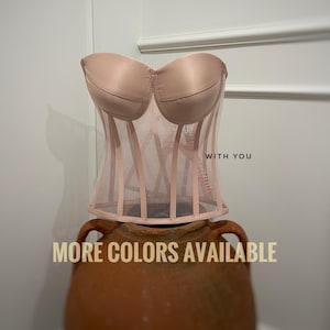 May include: A pink mesh corset with a satin bust. The corset has a boned structure and is displayed on a terracotta pot. The text 'WITH YOU' is visible on the corset. The text 'MORE COLORS AVAILABLE' is displayed below the corset.
