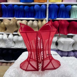May include: A red corset top featuring sheer mesh fabric, structured boning, and a front button closure. The garment displays the text WITH YOU on the front panel. It is positioned in front of shelves filled with various colored corset tops.