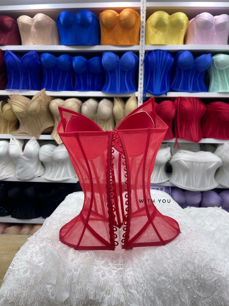 May include: A red corset top featuring sheer mesh fabric, structured boning, and a front button closure. The garment displays the text WITH YOU on the front panel. It is positioned in front of shelves filled with various colored corset tops.