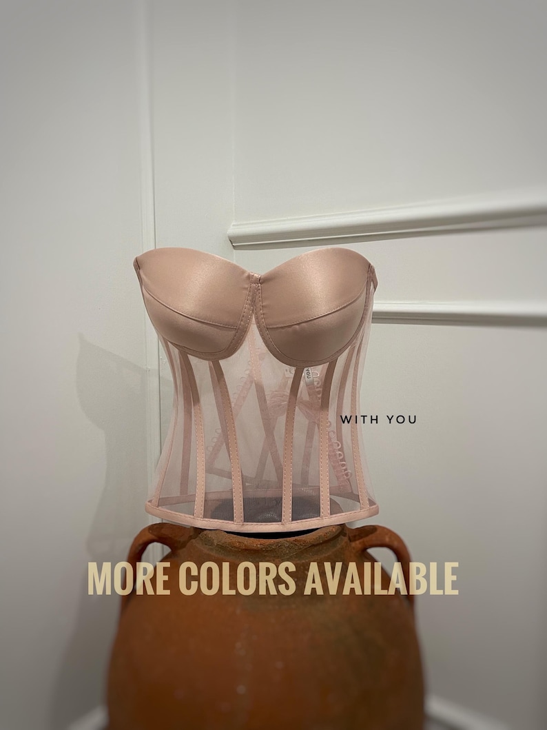 May include: A pink mesh corset with a satin bust. The corset has a boned structure and is displayed on a terracotta pot. The text 'WITH YOU' is visible on the corset. The text 'MORE COLORS AVAILABLE' is displayed below the corset.