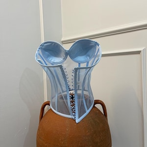 May include: A light blue sheer corset with satin cups and a lace-up front. The corset is on a terracotta-coloured pedestal.