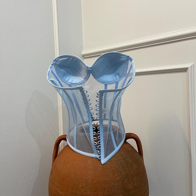 May include: A light blue sheer corset with satin cups and a lace-up front. The corset is on a terracotta-coloured pedestal.