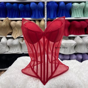 May include: A red corset top featuring a sheer mesh overlay, structured boning, and a sweetheart neckline. The garment has a pointed hemline and includes the text WITH YOU printed in black on the front right side of the bodice.