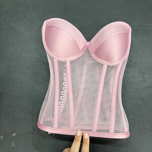 May include: A pink mesh corset with satin cups and boning. The corset has a strapless design and a hook and eye closure in the back.