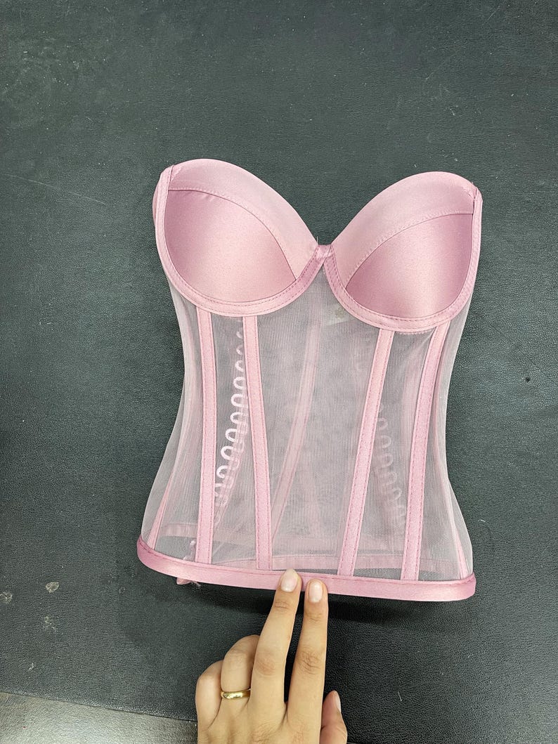 May include: A pink mesh corset with satin cups and boning. The corset has a strapless design and a hook and eye closure in the back.