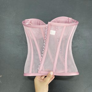 May include: A pink mesh corset with a satin trim and a row of metal eyelets down the center back.