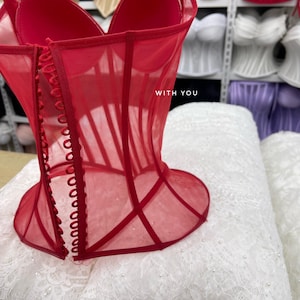 May include: A bright red corset top made of sheer mesh fabric with structured boning and a front button closure. The garment features a sweetheart neckline and the text WITH YOU printed in white on the front panel.