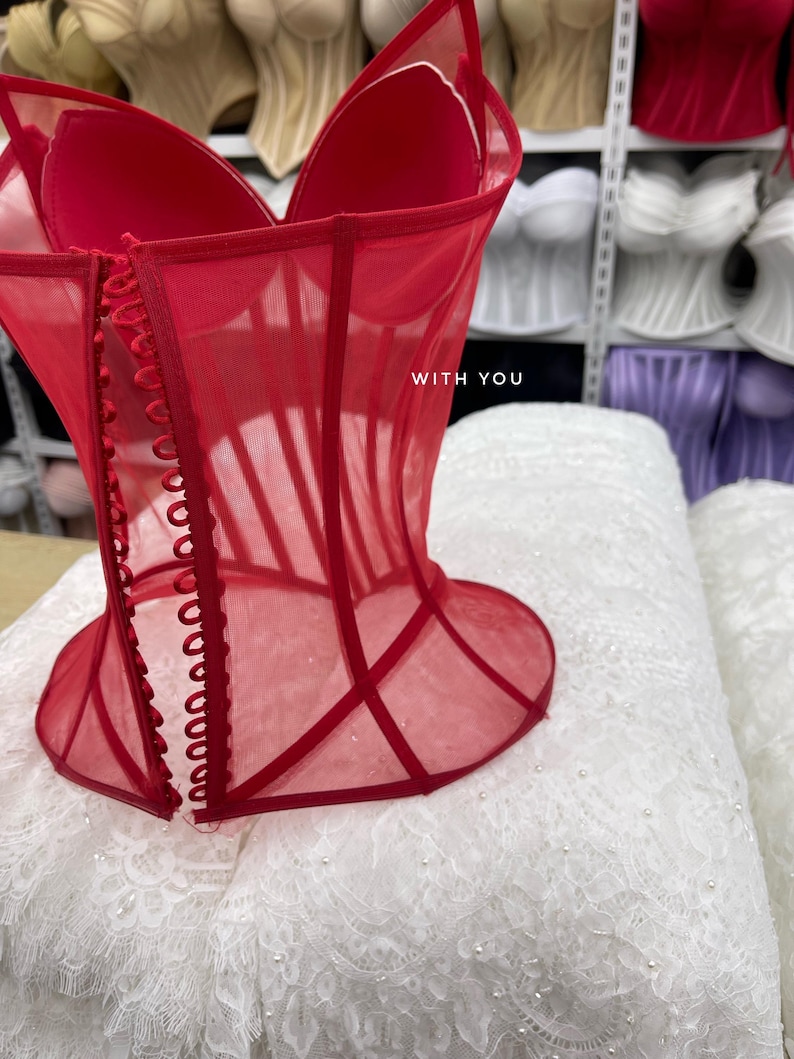 May include: A bright red corset top made of sheer mesh fabric with structured boning and a front button closure. The garment features a sweetheart neckline and the text WITH YOU printed in white on the front panel.