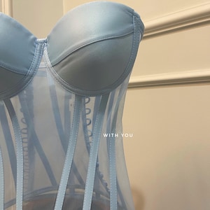 May include: A light blue strapless corset with a mesh panel and boning. The corset has a satin-like finish and features a row of buttons down the front. The text "WITH YOU" is printed on the front of the corset.