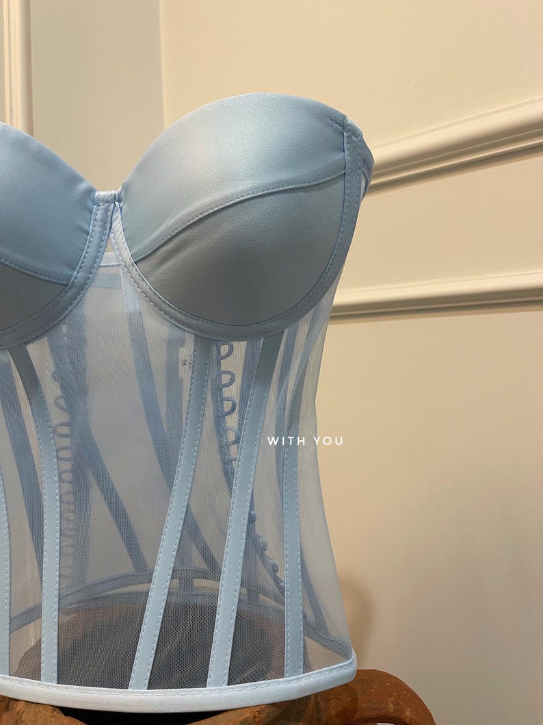 May include: A light blue strapless corset with a mesh panel and boning. The corset has a satin-like finish and features a row of buttons down the front. The text "WITH YOU" is printed on the front of the corset.