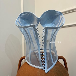 May include: A light blue mesh corset with a satin bodice and a row of silver eyelets down the centre back. The corset has a 'WITH YOU' label on the back.