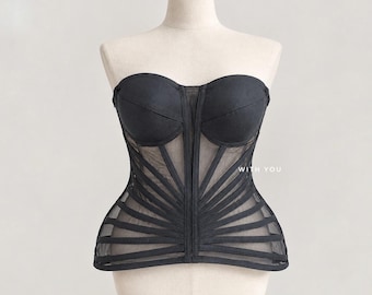 May include: A black strapless corset top featuring structured cups and a sheer mesh bodice with radiating black fabric strips. The garment includes a central vertical seam and the text WITH YOU printed in small white letters on the right side.