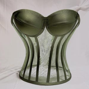 May include: A green mesh corset with a satin top and a boned, corseted bottom. The corset has a sweetheart neckline and is designed to cinch the waist.