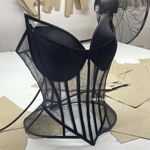May include: A black corset top featuring structured cups and sheer mesh panels with vertical boning details. The garment includes a pointed hemline and the text WITH YOU printed in white on the lower front section of the mesh fabric.