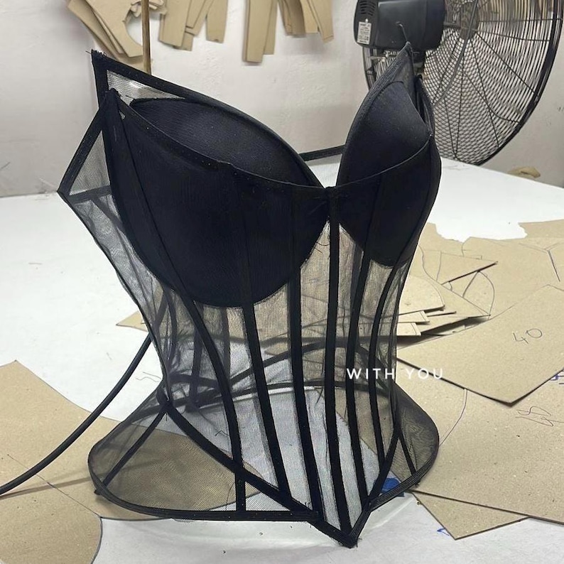 May include: A black corset top featuring structured cups and sheer mesh panels with vertical boning details. The garment includes a pointed hemline and the text WITH YOU printed in white on the lower front section of the mesh fabric.