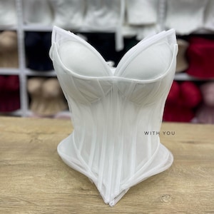 May include: A white corset top featuring structured cups, vertical boning channels, and a pointed hemline. The garment is crafted from sheer mesh fabric and includes the text WITH YOU printed in small black letters on the front right side.