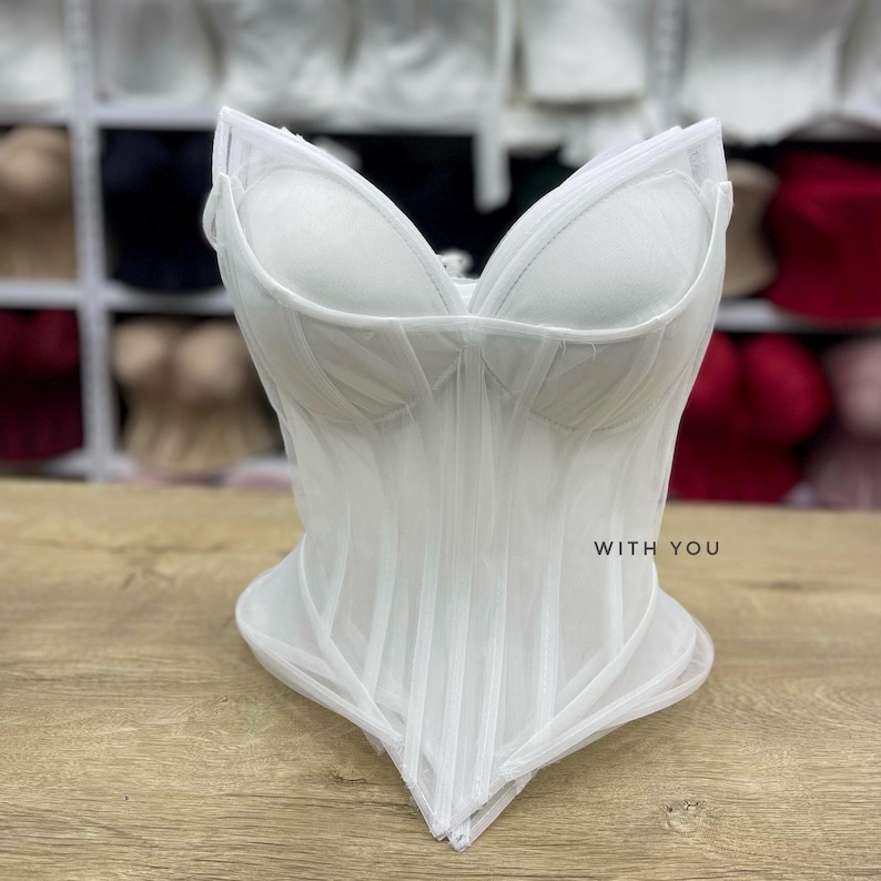 May include: A white corset top featuring structured cups, vertical boning channels, and a pointed hemline. The garment is crafted from sheer mesh fabric and includes the text WITH YOU printed in small black letters on the front right side.