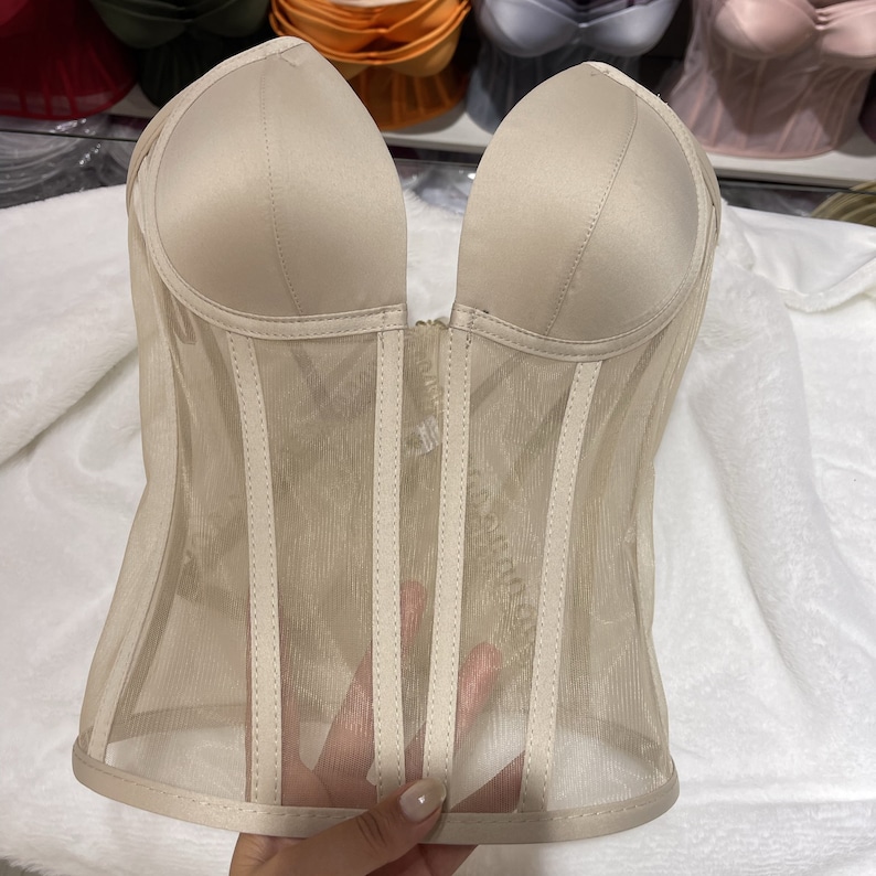 May include: A beige corset with a mesh overlay and a sweetheart neckline. The corset has boning and a hook and eye closure in the back.