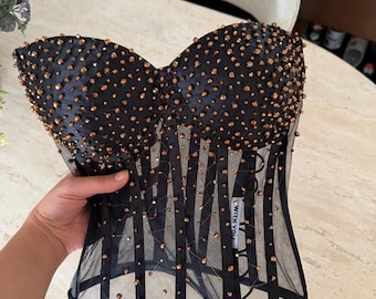 May include: A black corset top featuring a structured sweetheart neckline and sheer mesh panels. The garment is embellished with numerous small, metallic bronze-colored beads throughout. A white fabric label reads WITH YOU.