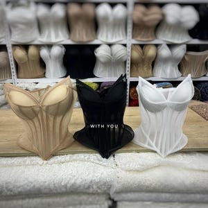 May include: Three structured corsets in beige, black, and white, each featuring molded cups and vertical boning. The black corset displays the text WITH YOU in white lettering on the front. These garments are displayed on a wooden surface.