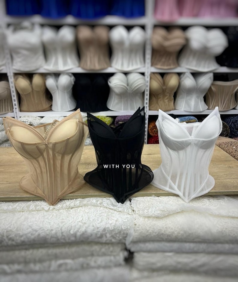 May include: Three structured corsets in beige, black, and white, each featuring molded cups and vertical boning. The black corset displays the text WITH YOU in white lettering on the front. These garments are displayed on a wooden surface.