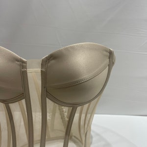 Pu&ograve; includere: Un corsetto in rete beige con una parte superiore in satin. Il corsetto ha una struttura a stecche e una chiusura a busk sul davanti.