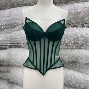 May include: A dark green corset top featuring sheer mesh panels, structured boning, and molded cups. The garment has a pointed hemline and includes the text WITH YOU printed in white on the right side of the bodice.