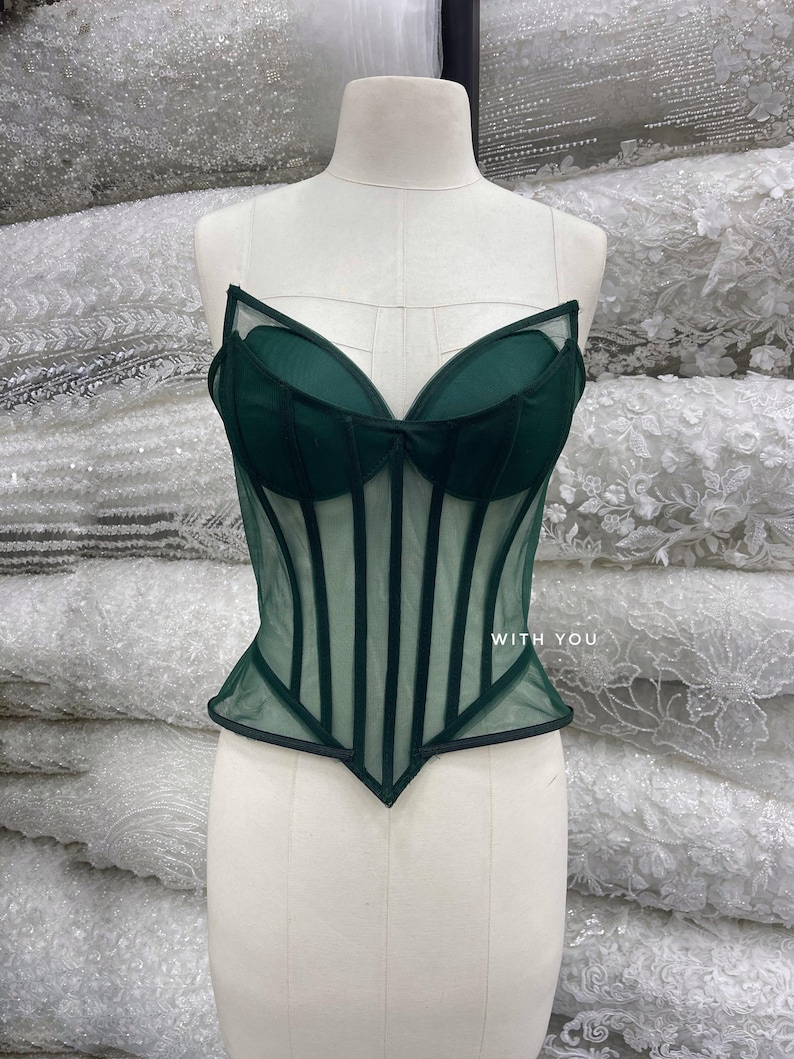 May include: A dark green corset top featuring sheer mesh panels, structured boning, and molded cups. The garment has a pointed hemline and includes the text WITH YOU printed in white on the right side of the bodice.