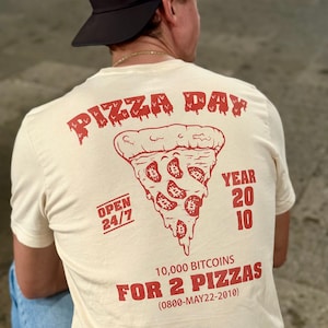 May include: A beige t-shirt with a red graphic of a pizza slice dripping with cheese. The pizza has bitcoin symbols on it. The text on the shirt reads "Pizza Day", "Open 24/7", "Year 2010", "10,000 Bitcoins for 2 Pizzas (0800-MAY22-2010)"
