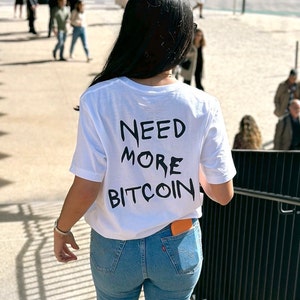 May include: A white t-shirt with the text "NEED MORE BITCOIN" printed on the back. The person wearing the shirt is walking down a set of stairs.