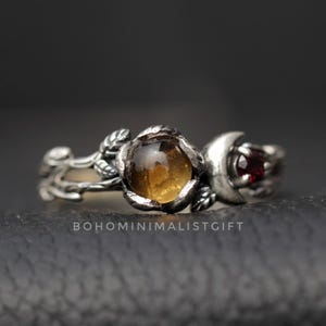 Könnte beinhalten: Ein silberner Ring mit floralem und himmlischem Design. Der Ring hat einen großen, runden, bernsteinfarbenen Edelstein und einen kleineren, roten Edelstein in Form eines Halbmonds. Das Band hat ein Blatt- und Rankenmotiv.