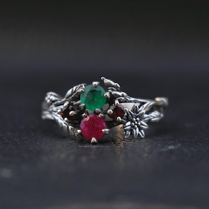 Peut inclure: Une bague en argent avec un motif floral, ornée d'une émeraude verte et d'un rubis rouge. La bague présente un design détaillé inspiré de la nature, avec des feuilles et une petite fleur. Les pierres précieuses sont rondes et serties au centre.
