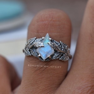 Peut inclure: Bague en argent ornée d'une pierre précieuse bleu clair au centre, surmontée d'une pointe. Le design de la bague est entouré de feuilles détaillées. Le texte "BOHOMINIMALISTGIFT" est visible en bas.