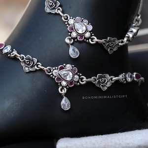 May include: Silver anklets with floral and teardrop accents. The anklets feature clear and ruby-colored gemstones. The design includes small flower details and a teardrop-shaped charm hanging from the center of the floral design.