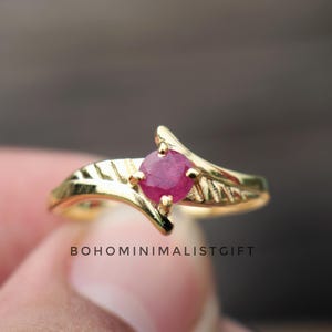 May include: A gold ring with a vivid red gemstone. The ring has a detailed leaf design on the band and a distinctive setting that holds the gemstone. The ring is a piece of jewellery. The text "BOHOMINIMALISTGIFT" is visible.