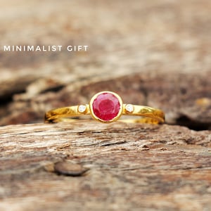 May include: A gold ring with a red gemstone and two smaller white gemstones. The ring is on a wooden surface. BOHO MINIMALIST GIFT