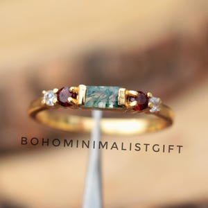 May include: A gold ring featuring a rectangular green stone, flanked by two red stones and two smaller white stones. The ring has a delicate band and the text "BOHOMINIMALISTGIFT" is visible.