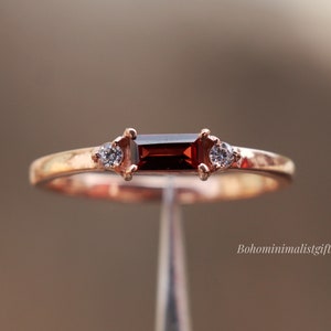 May include: A rose gold ring featuring a rectangular, dark red gemstone at the center, flanked by two smaller, clear gemstones. The ring is held up by a silver stand, with the text "Bohominimalistgift" visible in the background.