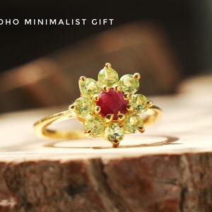 Op de afbeelding: Een gouden ring met een rode edelsteen in het midden, omringd door groene edelstenen, die samen een bloem vormen. BOHO MINIMALIST GIFT.