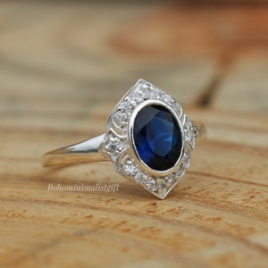 May include: A silver ring featuring a blue oval gemstone encircled by smaller clear gemstones. The ring boasts a delicate, intricate design.