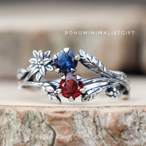 Op de afbeelding: Een zilveren ring met een delicaat bloemmotief met een blauwe en rode edelsteen. De ring ligt op een houten oppervlak.