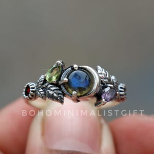 Puede incluir: Un anillo de plata con una piedra preciosa azul central, flanqueada por piedras más pequeñas verdes, rojas y moradas. El anillo presenta un diseño de hojas y el texto "BOHOMINIMALISTGIFT".