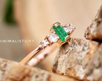 May include: A delicate gold ring with a green emerald gemstone and small clear diamonds. The ring is on a stack of wooden slices.