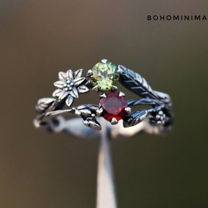 Peut inclure: Une bague en argent avec un motif floral et de feuilles, ornée d'une pierre verte et d'une pierre rouge. La bague porte le texte "BOHOMINIMALISTGIFT". C'est un bijou unique.