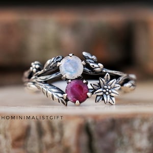 Puede incluir: Un anillo de plata con un delicado diseño floral con una piedra de luna y un rubí. El anillo está colocado sobre una superficie de madera.