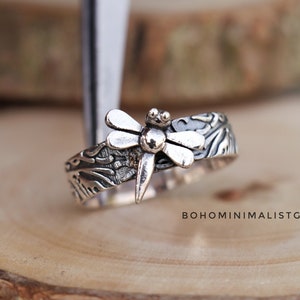 May include: Silver ring featuring a dragonfly design and a textured band. The band has a floral pattern.